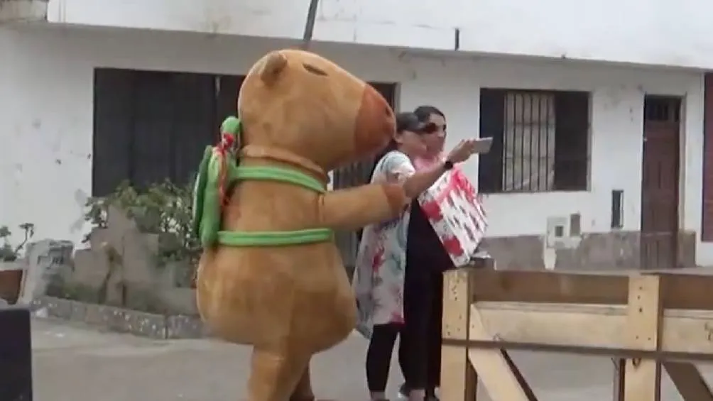 Un agente disfrazado de capibara, junto a una oficial de civil, simulan hacer una entrega especial con una caja de regalos. / RS
