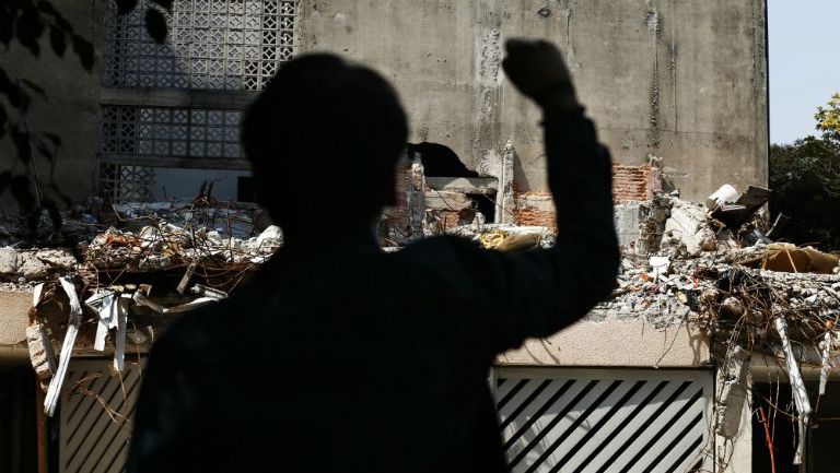 Silueta de un hombre con un puño en alto frente a un edificio colapsado en el que murieron varias personas